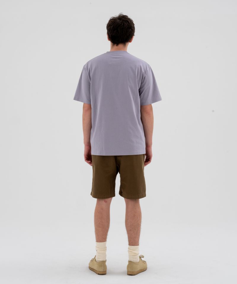 Back view of a model wearing a lavender Gramicci Half Dome Tee and brown shorts on a light-tone background.