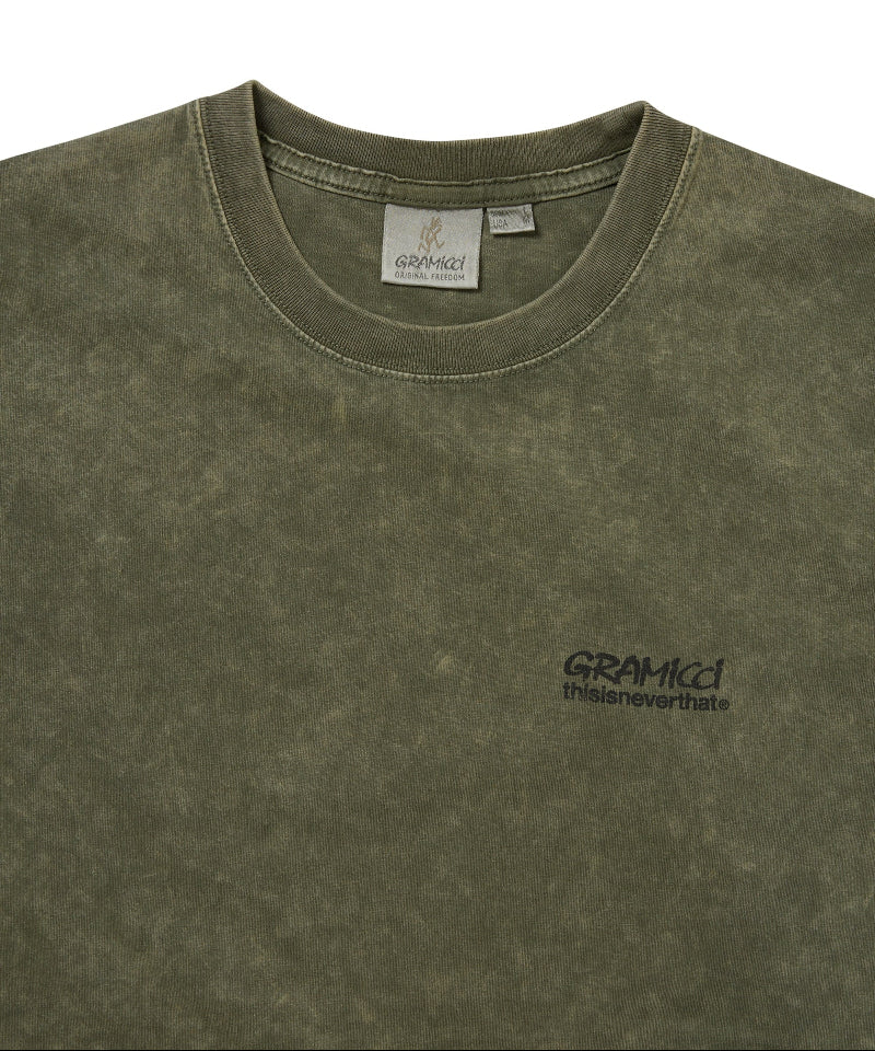 Close-up of an olive green mineral-washed t-shirt with a small black embroidered 'GRAMICCI' logo on the left chest, showing the inner neck label and fabric texture.
