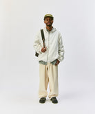 Model wearing a Gramicci light gray jacket, beige pants, and a cap on a light-tone background.